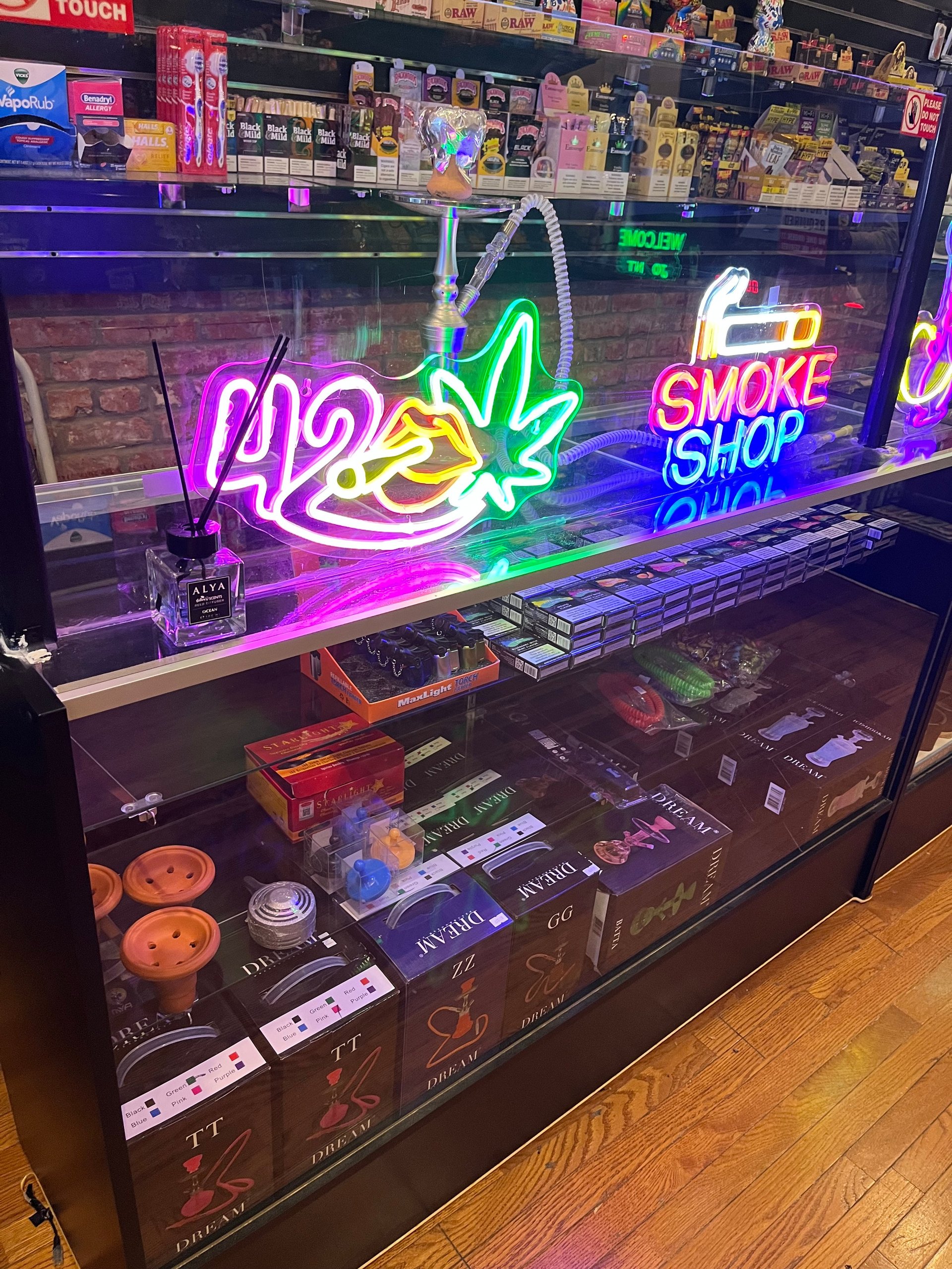 Neon sign display for a smoke shop with purple, green, and pink glowing text and cannabis leaf designs, merchandise visible below